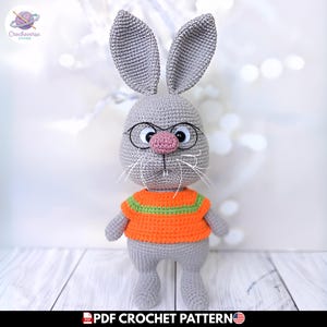 Puede incluir: Un muñeco de conejo tejido a ganchillo de color gris con orejas largas y erguidas, nariz rosa y gafas redondas negras. El conejo lleva un suéter naranja con una franja horizontal verde en el pecho. Bigotes de hilo blanco en la cara.