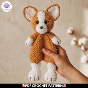 Op de afbeelding: Een gehaakt knuffeltje in de vorm van een hondje van bruin en wit garen. Het speelgoed heeft grote puntige oren, een witte snuit, zwarte veiligheidsoogjes en een klein zwart neusje. Het lichaam is zacht en getextureerd met witte pootjes.
