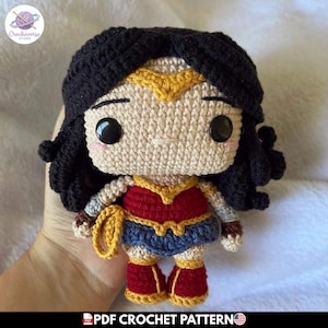 Puede incluir: Una muñeca de ganchillo con pelo negro, una tiara dorada y un conjunto rojo, azul y dorado. La figura tiene grandes ojos de botón negros, una pequeña boca bordada y mejillas sonrosadas. Un lazo amarillo está sujeto a la cintura.