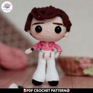 Puede incluir: Una muñeca de ganchillo hecha a mano con cabello castaño oscuro, grandes ojos negros y mejillas rosadas. La muñeca viste una camisa rosa de manga larga con cuello y pantalones blancos acampanados con detalles dorados. El texto PDF CROCHET PATTERN es visible.