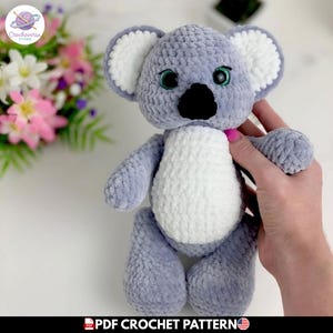 Puede incluir: Un peluche de koala hecho de hilo suave gris y blanco. El koala presenta una nariz negra grande, orejas redondas con centros blancos y ojos verdes. El cuerpo es redondo con una mancha blanca en el vientre. Es un patrón de ganchillo en PDF.