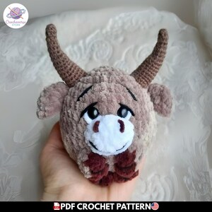 Puede incluir: Un juguete de ganchillo afelpado con forma de cabeza de vaca highland hecho de hilo marrón claro suave, con dos cuernos curvos marrón oscuro, orejas pequeñas, cejas bordadas en negro, ojos de plástico negros y un hocico blanco.