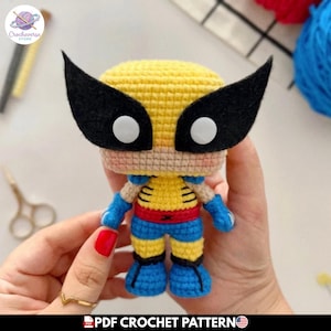 Puede incluir: Una pequeña figura amigurumi tejida a mano con cabeza amarilla, antifaces negros de fieltro y ojos circulares blancos. El cuerpo es amarillo con brazos y piernas azules, un cinturón rojo y detalles negros en un diseño estilizado y compacto.
