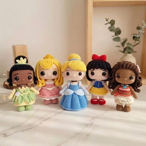May include: Five crocheted dolls representing various characters, each featuring large black eyes and distinct outfits. The dolls include a green dress with a leaf motif, a pink gown, a blue gown, a blue and yellow dress with a red bow, and a patterned top.