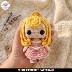 Puede incluir: Una pequeña muñeca de ganchillo con cabello de hilo amarillo brillante peinado con una corona, grandes ojos de plástico negro y un vestido rosa con ribete blanco. La muñeca está hecha con puntadas visibles y tiene tez beige claro.