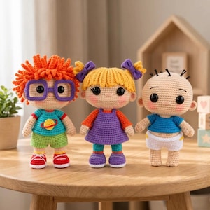 May include: Three crocheted dolls featuring characters with distinct appearances. The first has orange hair, purple glasses, a teal shirt with a planet graphic, and green shorts. The second has yellow hair with purple bows and a purple dress. The third is a baby in a blue shirt and white diaper.