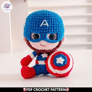 Puede incluir: Una muñeca de ganchillo hecha a mano de un superhéroe con cabeza grande, máscara azul con una letra A blanca y ojos negros grandes. La muñeca viste un traje azul y rojo con una estrella blanca en el pecho y sostiene un escudo redondo tricolor.