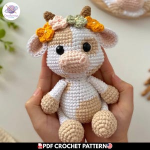 Puede incluir: Un pequeño peluche de vaca hecho a ganchillo con manchas blancas y beige, ojos de seguridad negros y dos pequeños cuernos marrones. La vaca está adornada con una corona de flores tejidas en tonos amarillo, rosa claro y verde salvia.