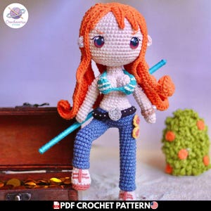 Puede incluir: Una muñeca de ganchillo con largo cabello naranja, grandes ojos oscuros y piel clara. La muñeca viste un top de bikini a rayas turquesa y blancas, pantalones azules con cinturón negro y zapatos blancos. Sostiene un bastón largo de color turquesa.