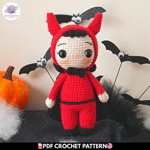 Puede incluir: Una muñeca tejida a mano con un disfraz de diablo rojo que incluye una capucha con dos cuernos, una pajarita negra y una capa negra. La muñeca tiene ojos negros grandes, piel clara y cabello negro. El texto PDF CROCHET PATTERN es visible.