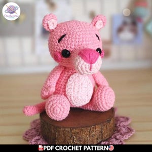 Puede incluir: Un animal de peluche tejido a ganchillo en color rosa con cabeza redondeada, orejas pequeñas, ojos de plástico negro y nariz rosa bordada. La figura tiene una mancha rosa más clara en el vientre, textura de hilo y una cola larga, sentado.