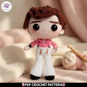 Puede incluir: Una muñeca de ganchillo con cabello castaño, ojos negros grandes y mejillas rosadas. La muñeca viste una camisa rosa de manga larga con cuello y pantalones blancos de talle alto con detalles naranjas. El texto PDF CROCHET PATTERN es visible.