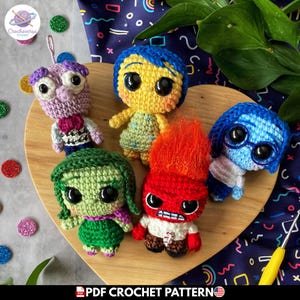 Puede incluir: Cinco muñecos de ganchillo, cada uno representando una emoción de la película "Del revés". Los muñecos son de color amarillo, azul, verde, rojo y morado. Se exhiben sobre una superficie de madera en forma de corazón. El texto "PDF Crochet Pattern" es visible.