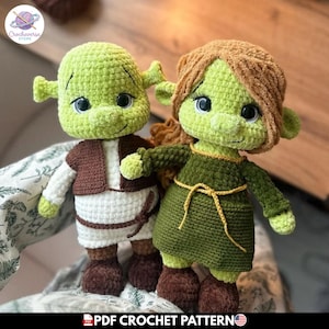 Puede incluir: Dos muñecos de ganchillo hechos a mano, uno que se asemeja a Shrek con un chaleco marrón y una camisa blanca, el otro la princesa Fiona con un vestido verde. Ambos muñecos son verdes con orejas grandes y caras sonrientes. Patrón de ganchillo PDF.