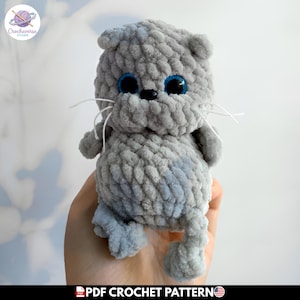 Puede incluir: Un pequeño juguete de peluche de gato gris hecho a mano con hilo grueso y suave. El gato presenta grandes ojos redondos de color azul brillante, una pequeña nariz negra y bigotes blancos finos a cada lado de la cara. Tiene un cuerpo redondeado y extremidades pequeñas.