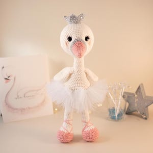 Puede incluir: Una muñeca de cisne tejida a ganchillo con cuerpo blanco, pico rosa suave y corona plateada. El cisne lleva un tutú de tul blanco y zapatillas de ballet tejidas en rosa con cordones cruzados. Ojos negros con pestañas largas y mejillas rosadas.