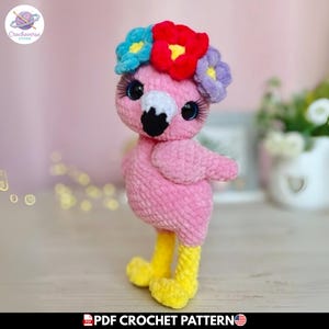 Puede incluir: Un flamenco de peluche rosa hecho de hilo suave con patas largas amarillas y un pico negro y blanco. El flamenco lleva una corona de flores tejidas a ganchillo en azul, rojo y morado. Tiene ojos grandes oscuros con pestañas negras largas. Texto PDF CROCHET PATTERN.