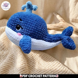 Puede incluir: Un juguete de peluche con forma de ballena hecho de hilo azul oscuro y blanco con una textura suave y esponjosa. La ballena tiene un pequeño chorro en la parte superior, ojos negros con reflejos blancos y una mejilla rosa. El texto PDF CROCHET PATTERN es visible.