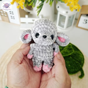 Può includere: Un piccolo peluche lavorato all'uncinetto a mano a forma di topo o animale simile. La figura è realizzata in morbido filato grigio testurizzato con grandi occhi neri rotondi, un piccolo naso nero e accenti rosa brillante all'interno delle orecchie.