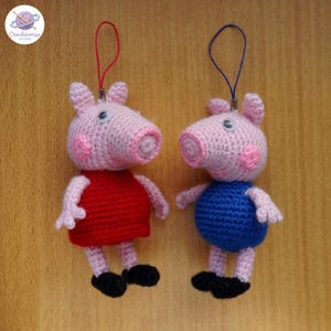 Pode incluir: Dois ornamentos de crochê feitos à mão em forma de porquinho com olhos móveis. Um porquinho usa um vestido vermelho com um cordão vermelho, enquanto o outro usa uma roupa azul com um cordão azul. Ambas as figuras têm corpos de fio rosa claro.
