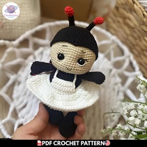 Puede incluir: Una muñeca de ganchillo con cabeza negra y dos antenas rematadas en esferas rojas, cara beige con ojos y boca bordados, y cuerpo negro vestido con un delantal blanco de ganchillo. La muñeca tiene brazos y piernas de color negro.