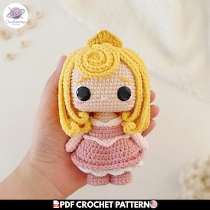 Puede incluir: Muñeca de ganchillo hecha a mano con cabello rubio y un vestido rosa. La muñeca tiene ojos negros grandes y lleva una corona amarilla. Patrón de ganchillo PDF.