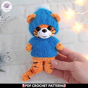 Puede incluir: Un pequeño peluche de tigre naranja con rayas negras, que lleva un suéter de punto azul brillante y un gorro azul a juego con un pompón blanco. El tigre tiene ojos de plástico negro y un hocico blanco. El texto inferior dice PDF CROCHET PATTERN.