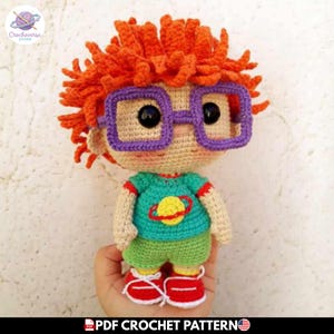 Puede incluir: Una muñeca de ganchillo con pelo naranja brillante, grandes gafas cuadradas moradas y ojos negros. La muñeca lleva una camiseta de color verde azulado con un gráfico de un planeta amarillo con un anillo rojo, pantalones cortos verdes.