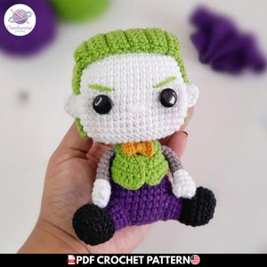 Puede incluir: Una pequeña muñeca de ganchillo con cara blanca, ojos de botón negros y pelo verde. La figura lleva un chaleco verde con pajarita naranja, pantalones morados, mangas blancas y zapatos negros. El texto PDF CROCHET PATTERN es visible abajo.