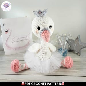 Puede incluir: Una muñeca cisne de ganchillo con cuerpo blanco, pico rosa y una corona gris con pequeñas gemas incrustadas. El cisne lleva un tutú de tul blanco y zapatillas de ballet rosas con detalles cruzados. La muñeca tiene ojos y pestañas negras.