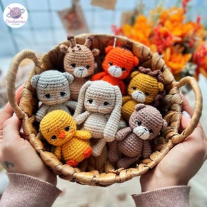 Puede incluir: Una cesta de mimbre tejida que contiene siete pequeñas figuras de animales de ganchillo, incluyendo un pollito amarillo, un conejo beige, un gato gris, una jirafa marrón, un tigre naranja, un león dorado y un oso marrón, con detalles bordados.