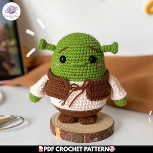 Puede incluir: Una muñeca de ganchillo verde con cabeza redonda, orejas pequeñas en forma de trompeta, ojos de cuentas negras y una sonrisa bordada. Viste un chaleco marrón sobre una camisa color crema y pantalones marrones. Texto PDF CROCHET PATTERN.