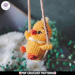 Puede incluir: Un pollito de ganchillo amarillo con un pequeño pico y patas naranjas, con un cuello blanco, suspendido por dos cuerdas de hilo beige. El pollito tiene ojos negros y mejillas rosadas. El texto PDF CROCHET PATTERN aparece abajo.