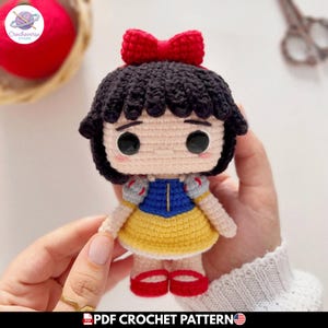 Puede incluir: Una muñeca de ganchillo que representa un personaje con cabello negro, un lazo rojo y un vestido azul y amarillo. La muñeca tiene ojos negros grandes, piel clara y zapatos rojos. El diseño incluye mangas abullonadas y una falda amarilla.