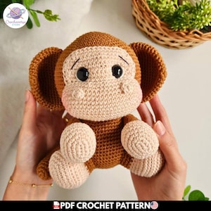 Puede incluir: Un juguete de mono tejido a ganchillo en marrón y beige con grandes orejas redondas, ojos de plástico negro y un hocico de color claro. La figura amigurumi tiene una superficie de hilo texturizado y un patrón de punto denso en el cuerpo.