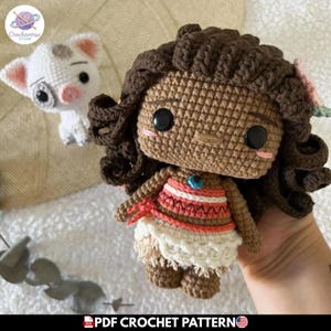 Puede incluir: Una muñeca de ganchillo con pelo rizado castaño oscuro, un accesorio de flor rosa y ojos de botón negros. La muñeca lleva un top estampado blanco y naranja con una falda blanca. A su lado hay un pequeño cerdito de ganchillo blanco.