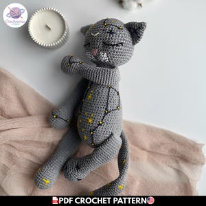 Puede incluir: Un juguete de peluche de gato tejido a ganchillo en color gris con constelaciones de estrellas amarillas bordadas y líneas negras en el cuerpo. El gato tiene los ojos cerrados, una nariz rosa y bigotes blancos bordados. Junto a una vela blanca.