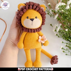 Puede incluir: Muñeco de león tejido a mano en crochet, en tonos amarillo y marrón, sosteniendo una flor rosa. El león tiene una melena marrón y una cara color crema. El texto "PDF CROCHET PATTERN" está en la parte inferior.