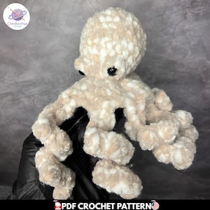 Puede incluir: Un pulpo de peluche hecho de hilo suave y jaspeado en tonos beige y blanco. La criatura presenta una cabeza redondeada, ocho tentáculos rizados y dos pequeños ojos de seguridad negros y brillantes. La textura es esponjosa y gruesa al tacto.