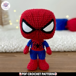 Puede incluir: Una muñeca tejida a mano con una cabeza roja grande, ojos blancos y negros, torso rojo con un emblema de araña negra, brazos y piernas azules, y pies rojos. La figura está hecha con puntadas visibles y tiene un diseño de cabeza grande.