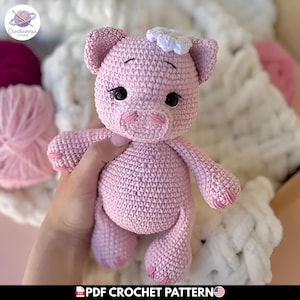 Puede incluir: Un peluche de cerdo tejido a ganchillo en color rosa, con una pequeña flor blanca decorativa en la cabeza, ojos de seguridad negros y detalles faciales bordados. El juguete tiene un cuerpo redondeado, extremidades cortas y un hocico definido.