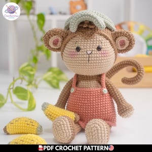 Puede incluir: Muñeco de mono tejido a ganchillo con cuerpo marrón claro, peto rosa y gorro de hoja de plátano verde. El mono sostiene un plátano amarillo y tiene mejillas rosadas. El muñeco está hecho a mano y parece un juguete blando.