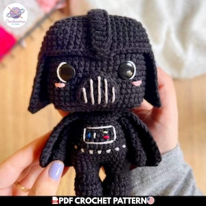 Puede incluir: Una muñeca de ganchillo negra con un casco grande, una rejilla bucal cosida en blanco y dos ojos negros redondos. La placa del pecho es rectangular con pequeños detalles bordados en rojo, azul y verde. El texto PDF CROCHET PATTERN es visible.