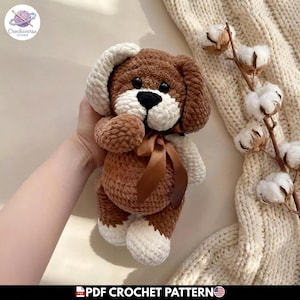 Puede incluir: Un juguete de peluche para perro hecho a ganchillo en color marrón y crema, con un acabado de hilo suave y texturizado, ojos de plástico negro, nariz bordada en negro y una cinta de satén marrón atada al cuello. Tiene orejas caídas.