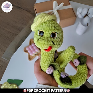 Peut inclure: Un serpent en crochet vert clair avec un petit chapeau assorti, de grands yeux sombres aux reflets dorés et une langue rouge. Le corps est enroulé et orné de petites fleurs roses et de feuilles vert foncé fixées sur la surface en laine texturée.