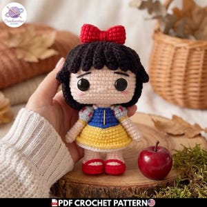 Puede incluir: Una muñeca de ganchillo con pelo negro y un lazo rojo, corpiño azul, falda amarilla y zapatos rojos. La muñeca tiene grandes ojos negros y piel clara. Una pequeña manzana roja está al lado. El texto PDF CROCHET PATTERN es visible.