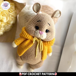 May include: A tan crocheted rabbit toy wearing a bright yellow knitted cardigan with a tie front. The rabbit has dark eyes, a pink stitched nose, and long ears. The text at the bottom reads PDF CROCHET PATTERN with a small United States flag icon.