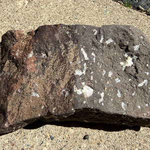 Peut inclure: Une grosse roche de forme irrégulière avec une surface rugueuse et texturée. La roche est principalement gris foncé avec des taches brun rougeâtre et des inclusions cristallines blanches. La pierre est posée sur une surface en béton.