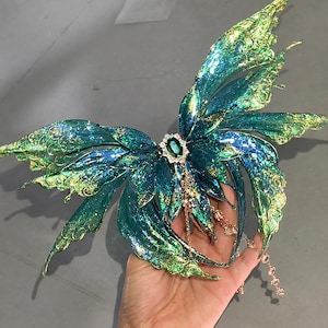 May include: A decorative butterfly-shaped accessory featuring iridescent teal and green wings with gold-toned filigree edges. The center is adorned with an oval emerald-colored gemstone surrounded by small clear crystals and dangling gold chains.