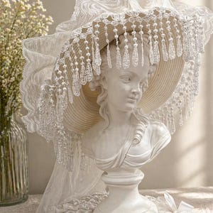 Puede incluir: Un busto blanco de mujer con un sombrero de ala ancha con encaje y flecos de cuentas. El sombrero está adornado con encaje delicado y un velo fluido. El busto está sobre un pedestal, con un fondo suave y neutro.